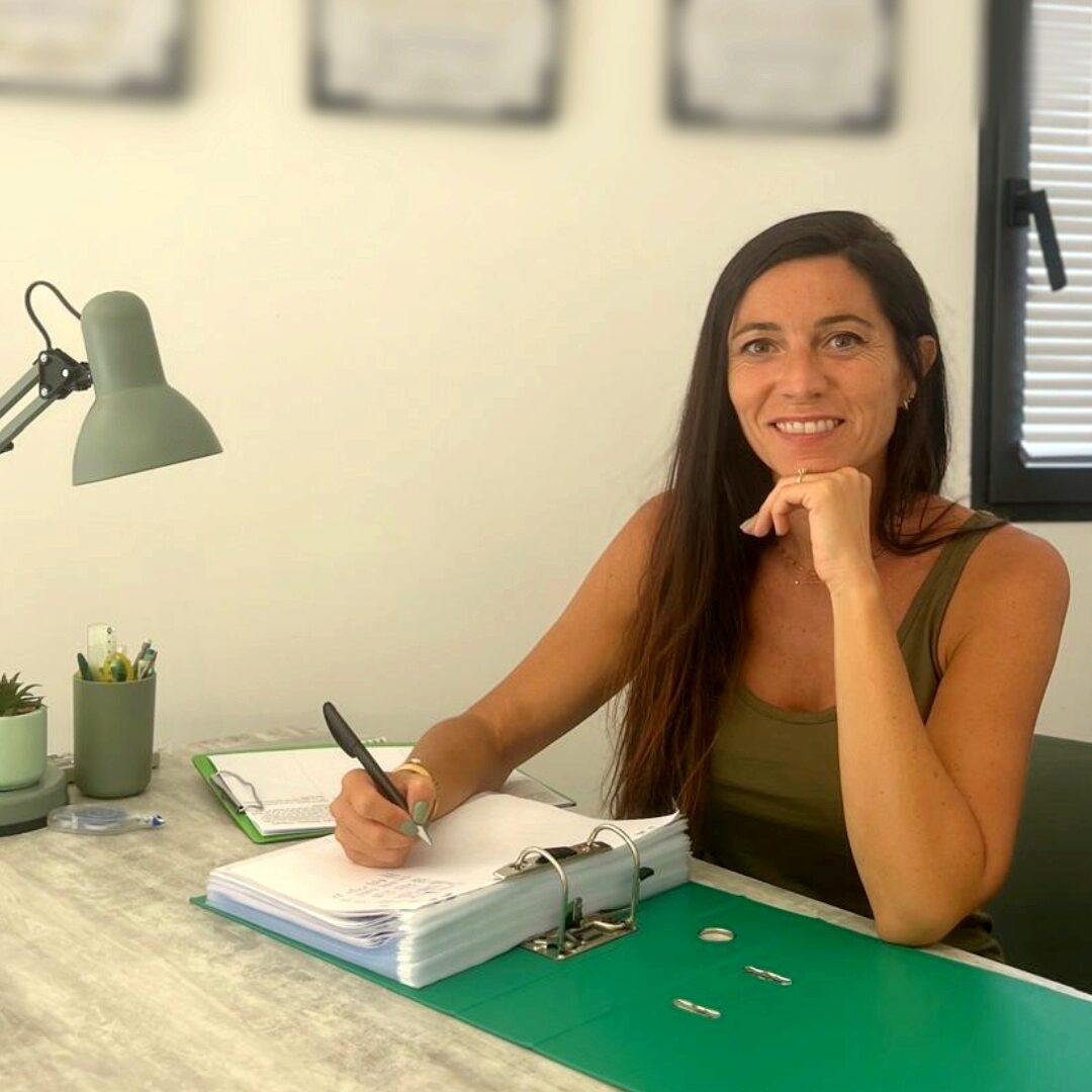 L'image montre une femme assise à un bureau, souriante, tenant un stylo dans sa main droite alors qu'elle semble prendre des notes dans un classeur ouvert. Elle est entourée d'objets de bureau, dont une lampe et un pot de crayons. En arrière-plan, des diplômes sont accrochés au mur. L'image dégage une ambiance professionnelle et accueillante, mettant en avant une personne concentrée et prête à recevoir des clients ou patients.